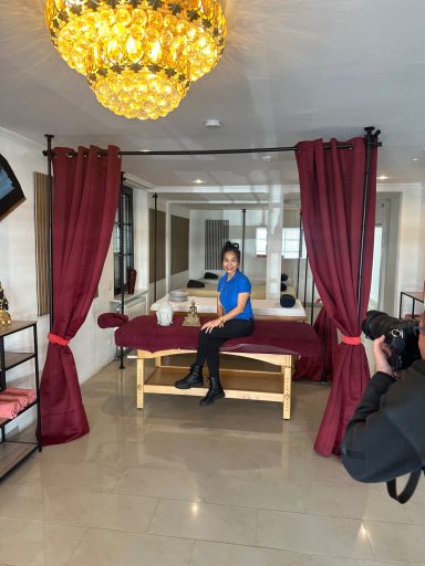 Frau in blauer Bluse sitzt auf einer Liege, umgeben von roten Vorhängen und einer Deckenlampe.